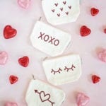 Four white canvas pouches with red rhinestone assorted designs. Lying on a pink background with pink and red glitter hearts scattered around
