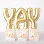 YAY written in gold balloon text figurine on top of three takeout containers with conversation hearts stickers on the front